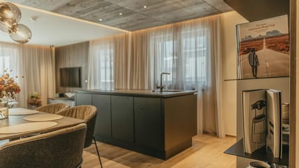 Moderner Wohnraum mit Esstisch, bequemen Stühlen, Kücheninsel und Bücherregal, helle Vorhänge, warme Holzdecke.
