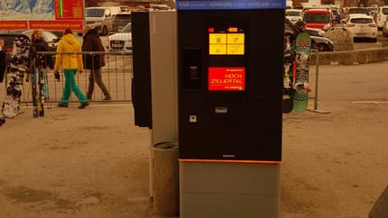 Ski pass machine with screen, in a parking lot with mountains in the background.
