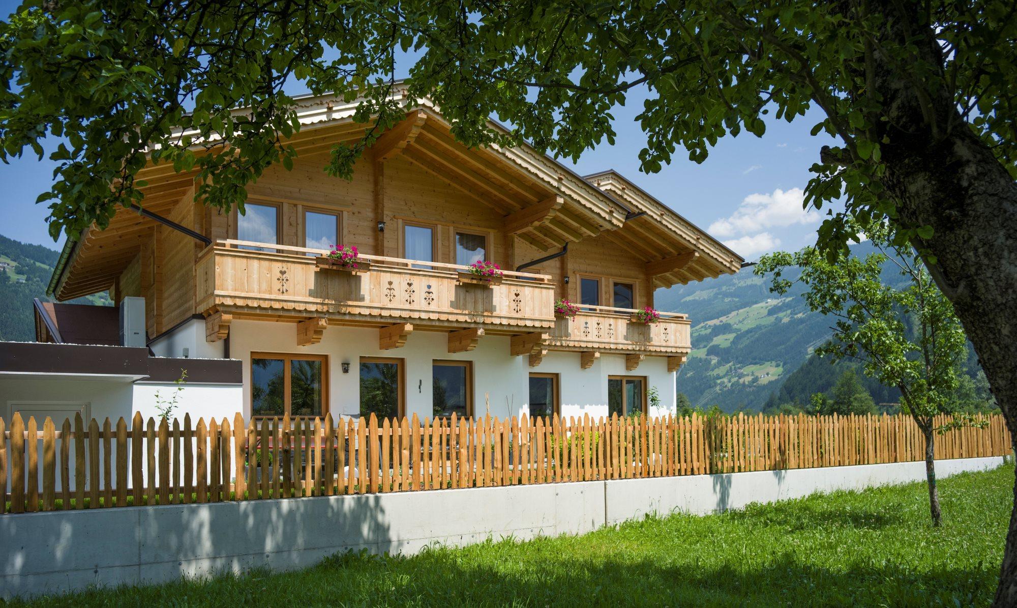 Häuser mit Holzfassade und Balkon, umgeben von Bäumen und Garten, mit Bergen im Hintergrund.