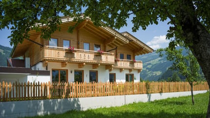 Häuser mit Holzfassade und Balkon, umgeben von Bäumen und Garten, mit Bergen im Hintergrund.