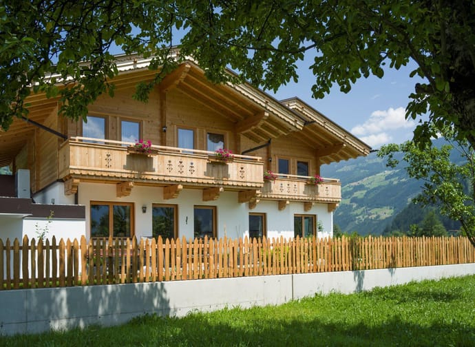 A wooden house with balconies and flowers, surrounded by trees and a garden, with mountains in the background.