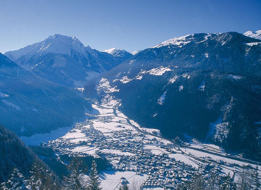 Snow-covered mountain village in a valley, surrounded by tall, snowy peaks and dense forests.