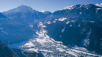 Snow-covered mountain village in a valley, surrounded by tall, snowy peaks and dense forests.