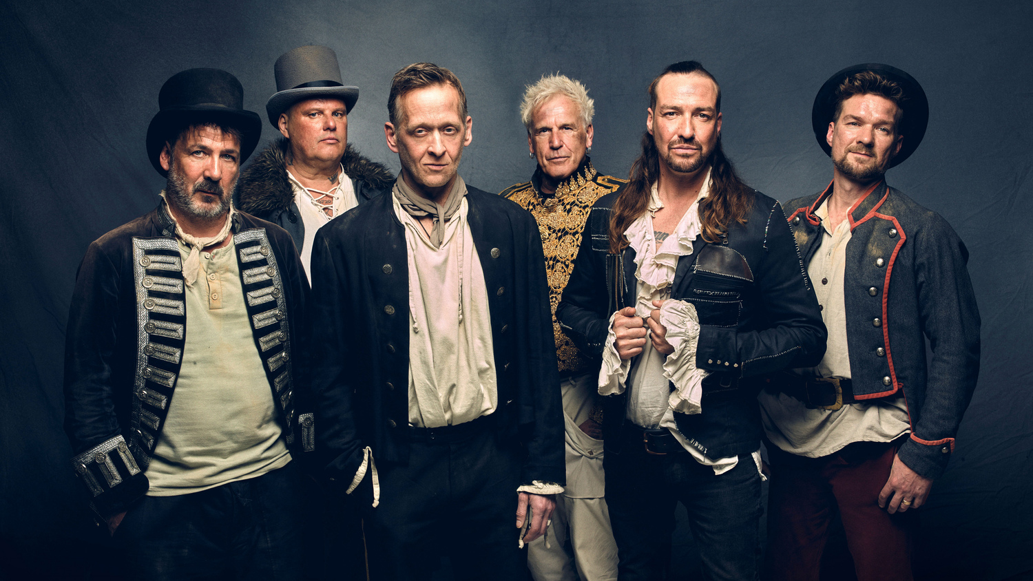 Six men in medieval-inspired outfits stand side by side against a dark background. They wear different hats, jackets, and shirts; some have long hair or beards. The group looks like a folk/medieval ensemble.