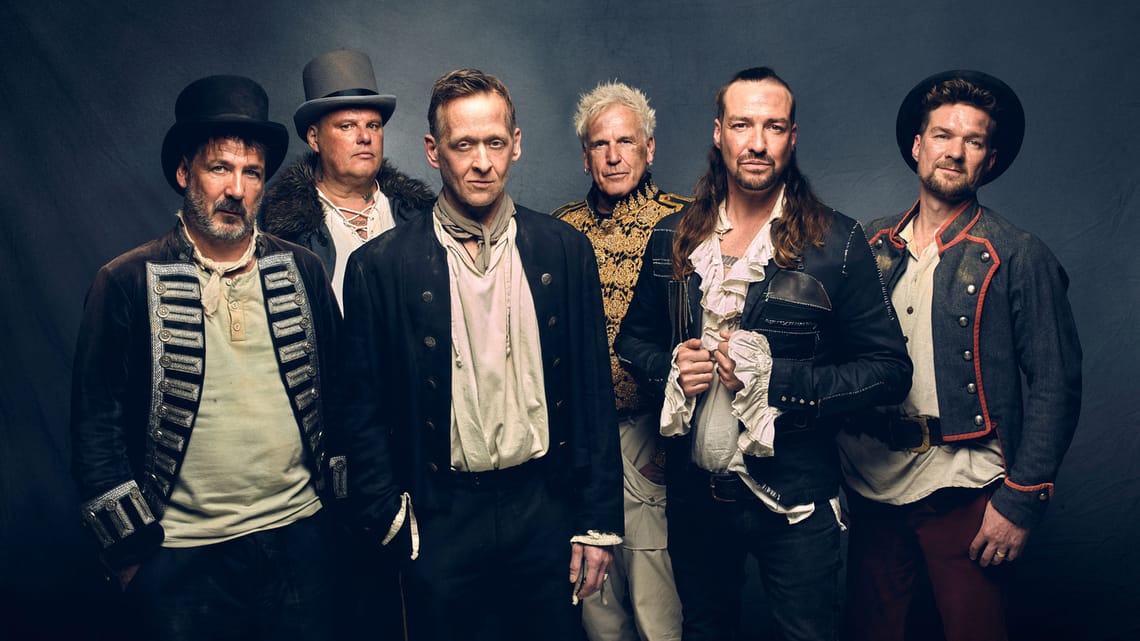 Inextremo die Gruppe beim Full Metal Mayrhofen 2026 Sechs Männer in mittelalterlich inspirierten Outfits stehen nebeneinander vor dunklem Hintergrund. Verschiedene Hüte, Jacken und Hemden; teils lange Haare oder Bart. Die Gruppe wirkt wie ein Folk-/Mittelalter-Ensemble.