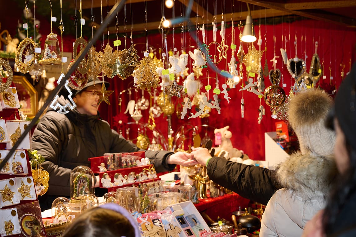Mayrhofner Advent - Verkaufsstände in Mayrhofen Auf dem Adventmarkt reicht ein Verkäufer in einer dicken Jacke und mit Brille einer Kundin einen Gegenstand über einen Tresen voller glitzernder Weihnachtsdeko. Über dem Stand hängen unzählige goldene und weiße Christbaumschmuckstücke.