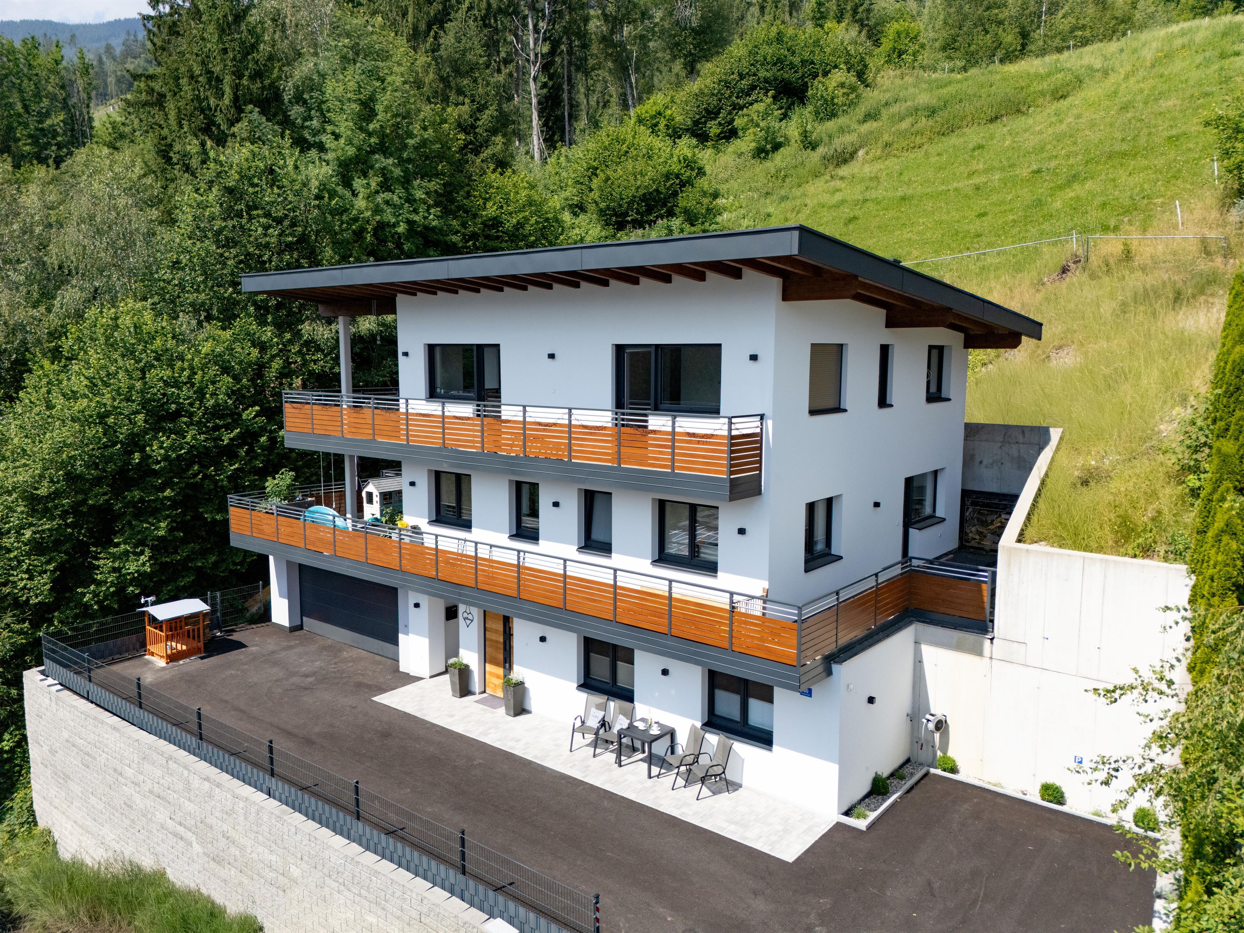 Modern house on a hill with balcony, surrounded by trees and greenery, with mountains in the background.