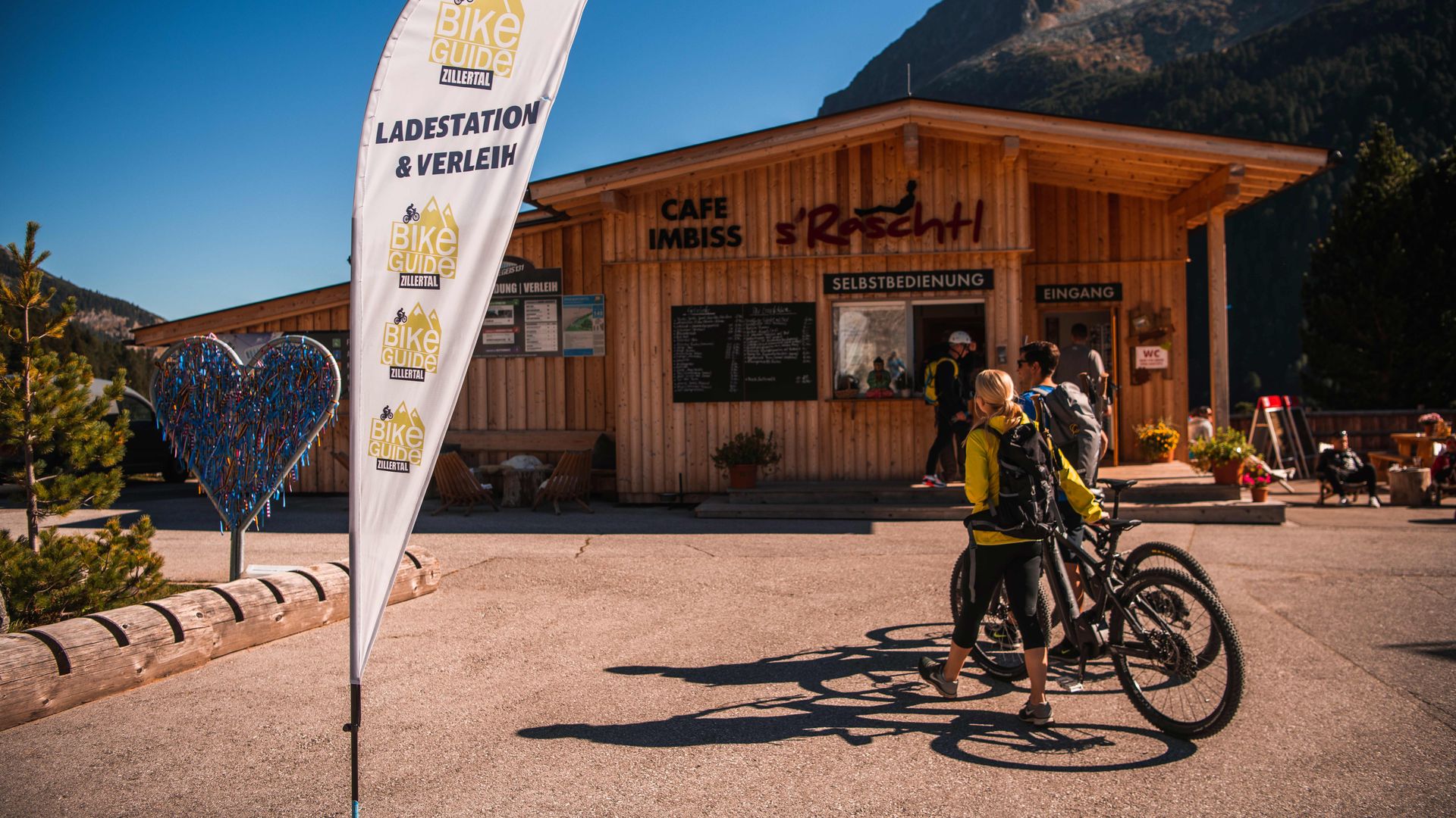 Zwei E-Biker vor einer E-Bike-Ladestation in der Ferienregion Mayrhofen-Hippach an einem sonnigen Tag – mit Infostand und Gebirgskulisse im Hintergrund.