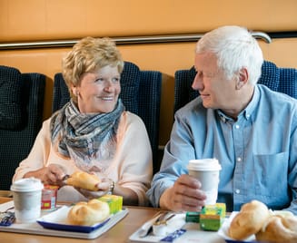 Älteres Ehepaar genießt gemeinsam Frühstück im Nightjet-Speisewagen. Sie sitzen sich an einem Tisch gegenüber, trinken Kaffee und essen Gebäck. Blaue Polstersitze und warme Atmosphäre.