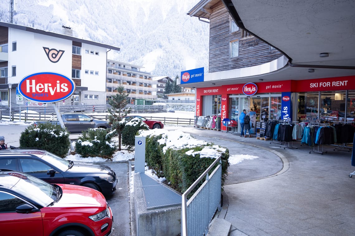 A Hervis Sports store in a winter mountain landscape. Jackets and ski clothing hang in front of the entrance, and signs indicate ski rental. Cars are parked in front, and snow lies on the hedges and at the roadside.