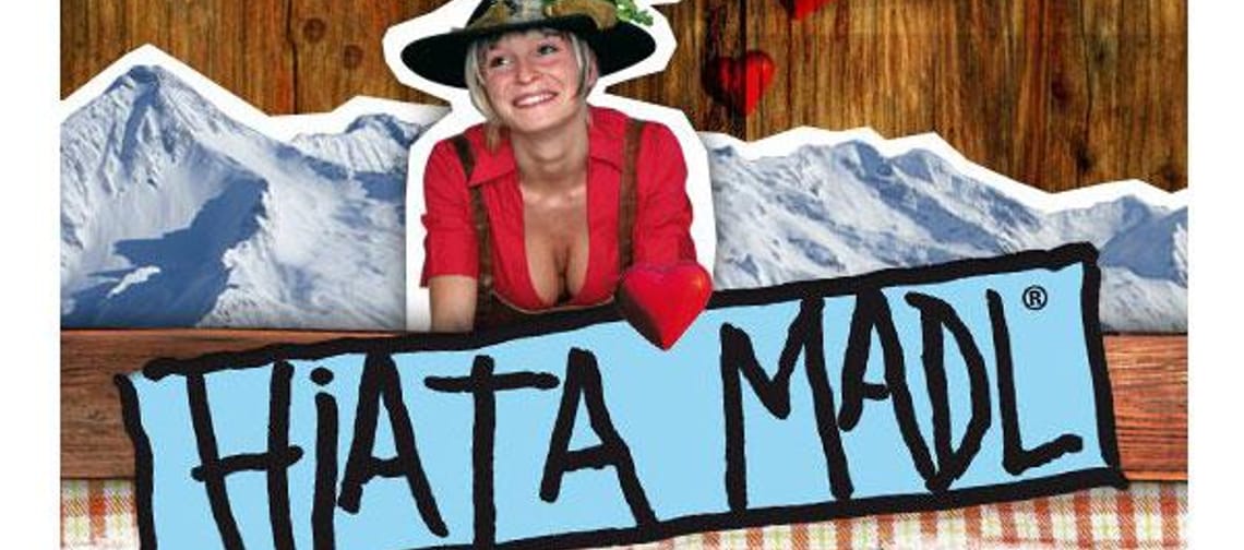 Das Logo vom Hiata Madl zeigt den Schriftzug „Hiatamadl“ in schwarzer Schrift mit einem blauen Rahmen auf einem Holz-Hintergrund. Dahinter sind stilisierte Berge und eine Frau mit Tirolerhut zu sehen – verziert mit roten Herzen. Das Design wirkt tradition