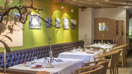 Ein gemütlicher Restaurantraum mit grüner Wand, Fotos, Tischen mit weißen Tischdecken und Stühlen.