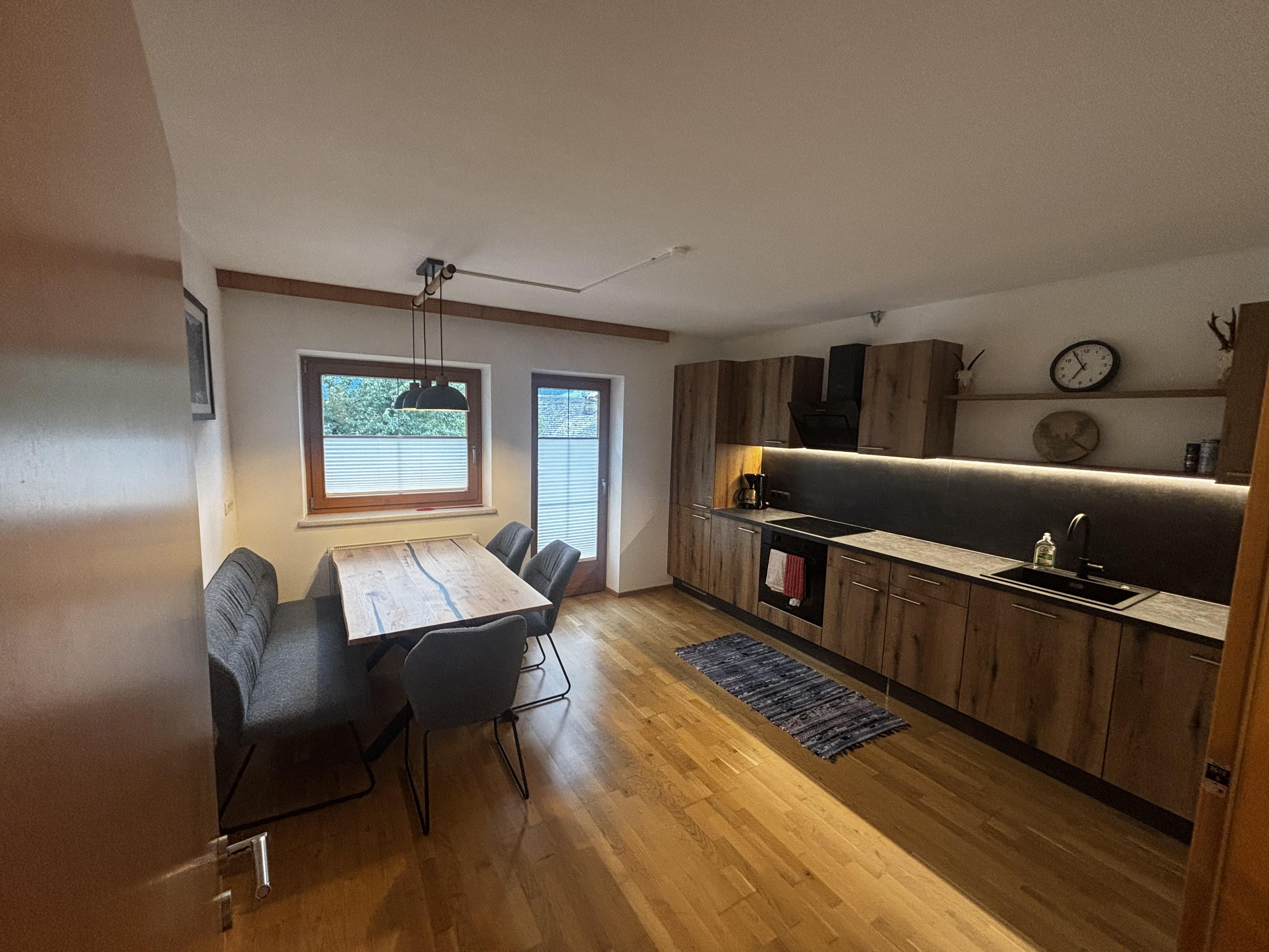 Küche mit Holzschränken, Esstisch, Stühlen, Fenster und modernen Geräten, helle und warme Atmosphäre.