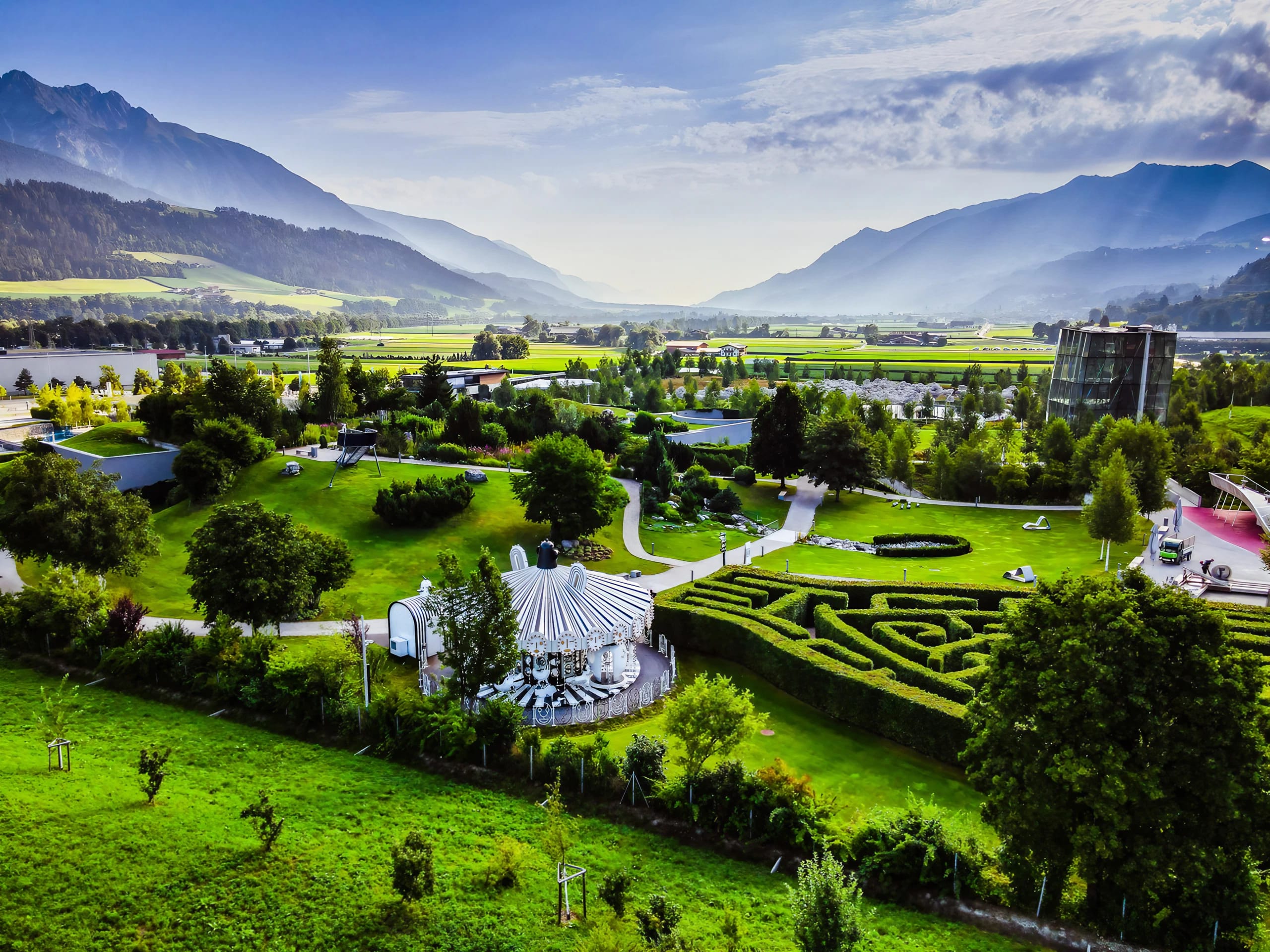 A beautiful garden with a carousel, surrounded by green trees and colorful flowers, with mountains in the background.
