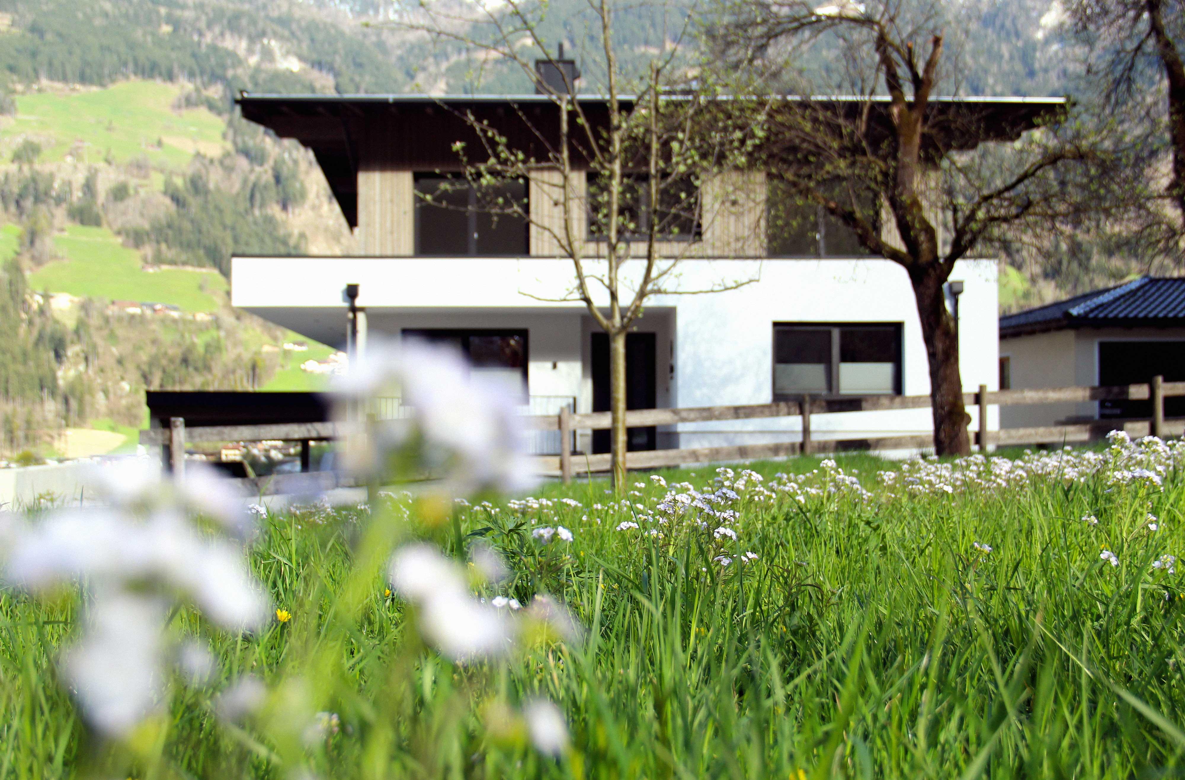 Modernes Haus mit weißen Wänden, große Fenster und umliegender Garten mit Bäumen und Blumen.