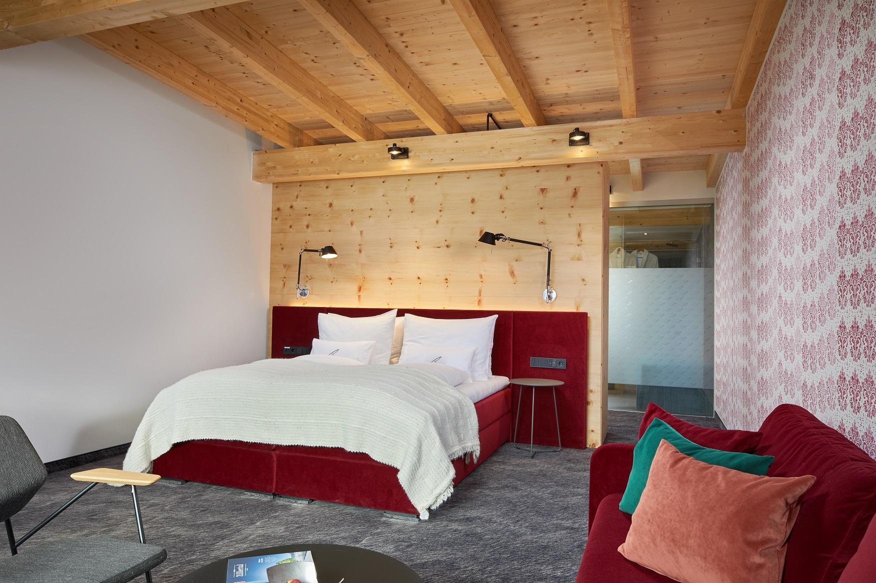 Modernes Hotelzimmer mit rotem Bett, Holzdecke und Wandlampen, gemütliche Atmosphäre, Sofa mit Kissen, Blick auf die Tür.