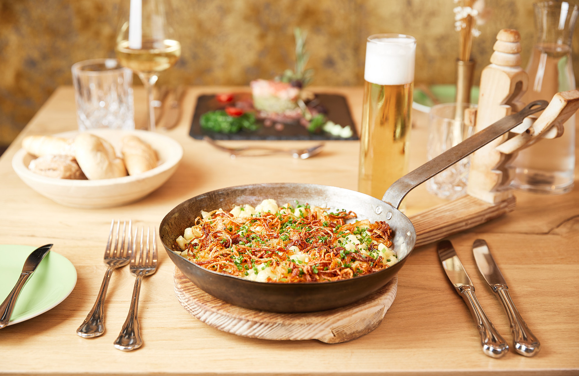 Käsespätzle in einer Eisenpfanne mit knusprigen Röstzwiebeln und Schnittlauch auf einem Holztisch; im Hintergrund Bier, Brot und ein Glas Weißwein.