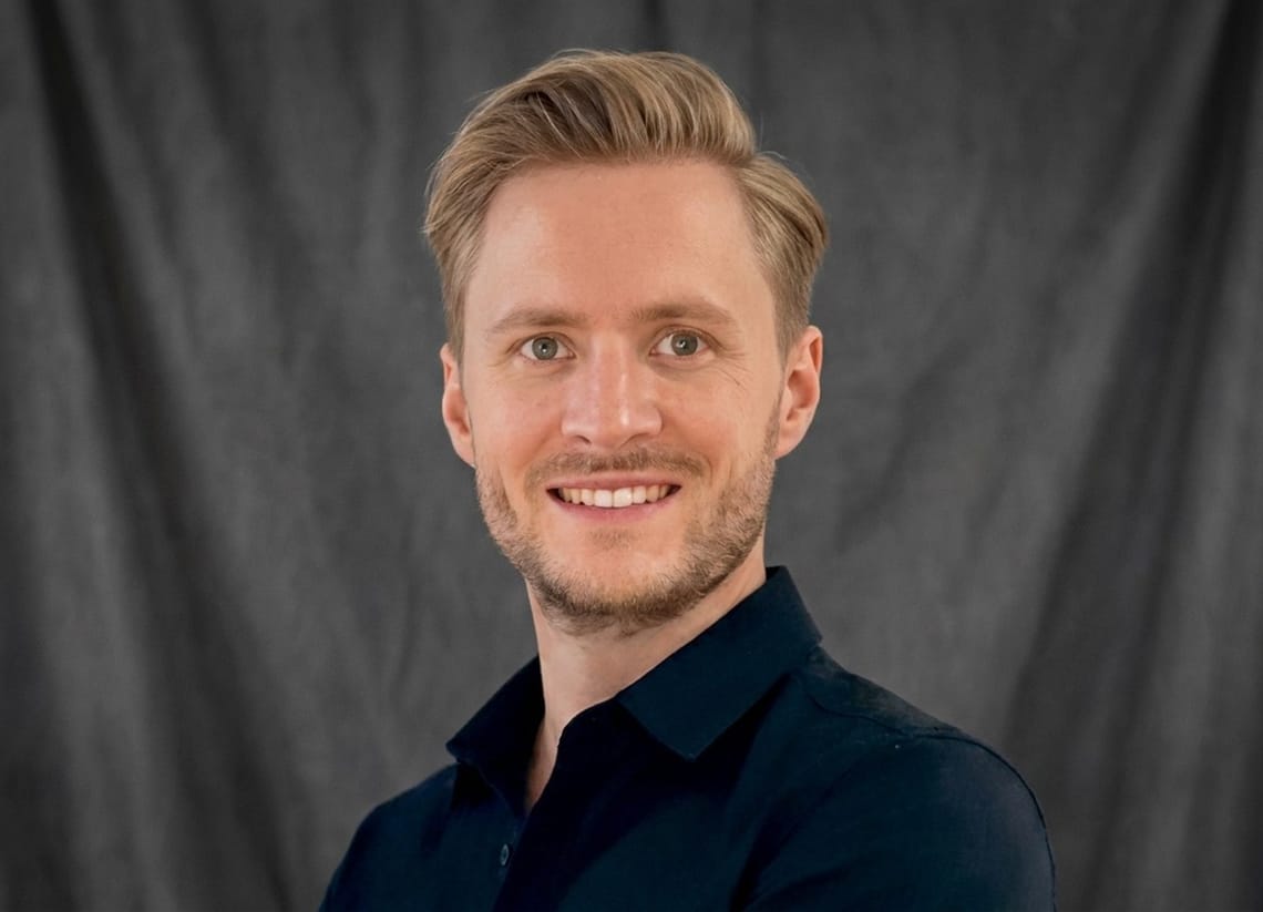 Niklas Kramer – Press Officer TVB Mayrhofen-Hippach Portrait photo of Niklas Kramer, press officer of TVB Mayrhofen: man with short brown hair, a well-groomed beard and light eyes in a dark navy shirt, smiling slightly against a dark grey fabric background.