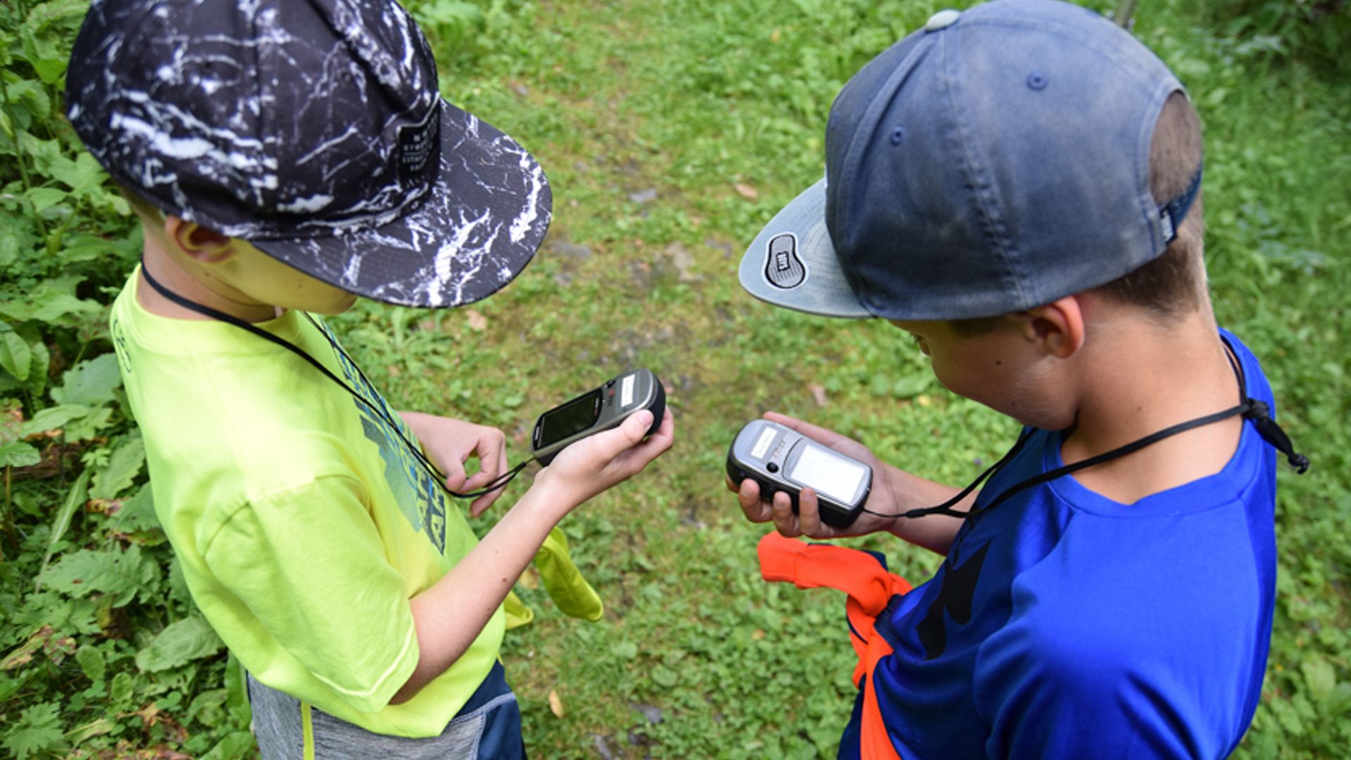 Two children wearing caps stand on a forest path in Ginzling, Zillertal. They hold a GPS device and a compass while searching for a geocache.