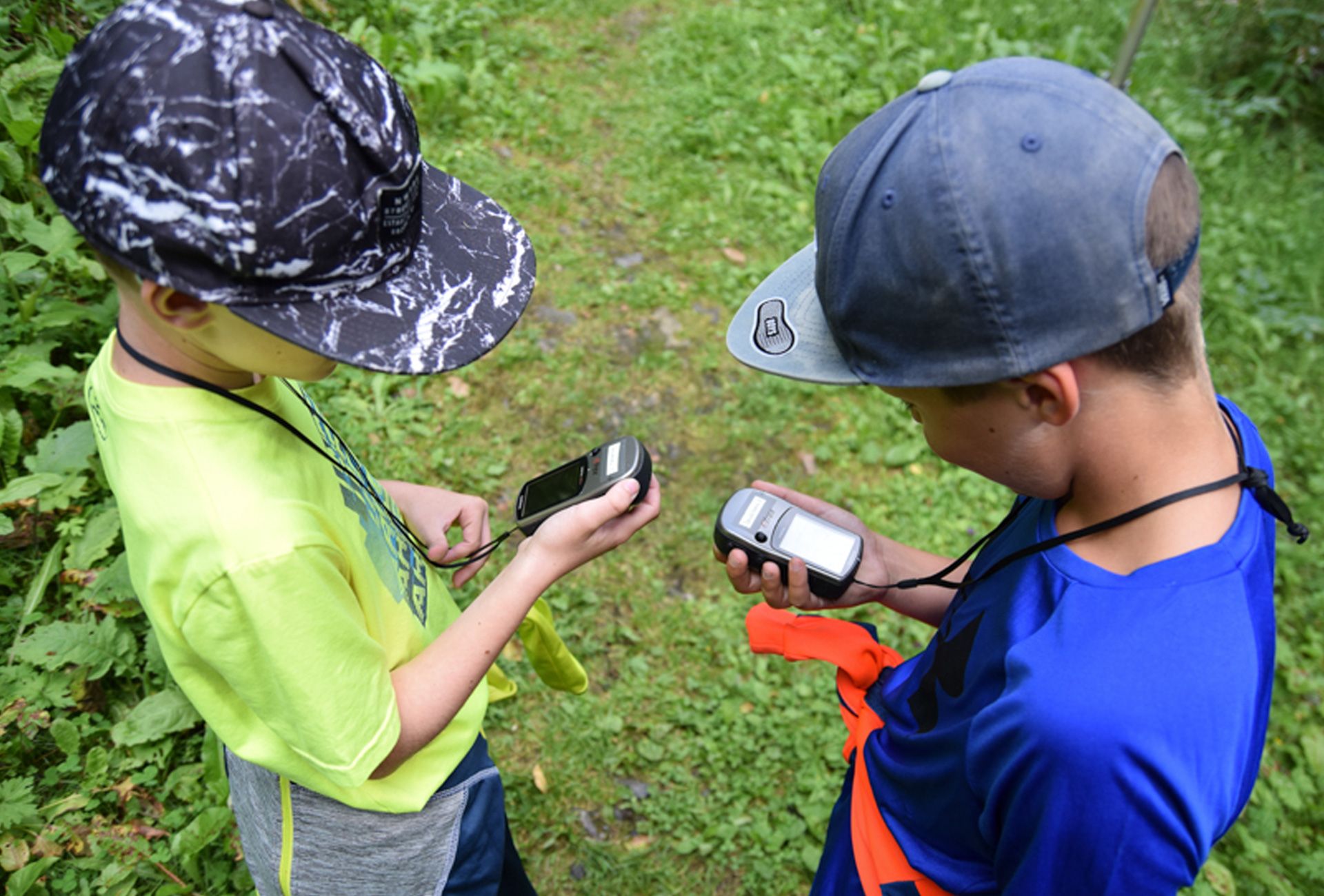 Two children wearing caps stand on a forest path in Ginzling, Zillertal. They hold a GPS device and a compass while searching for a geocache.