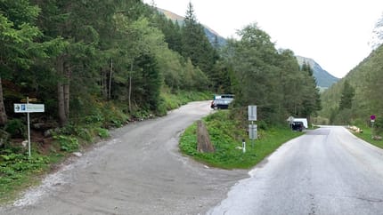 A narrow dirt road in a forested mountain area with parked cars and signs.
