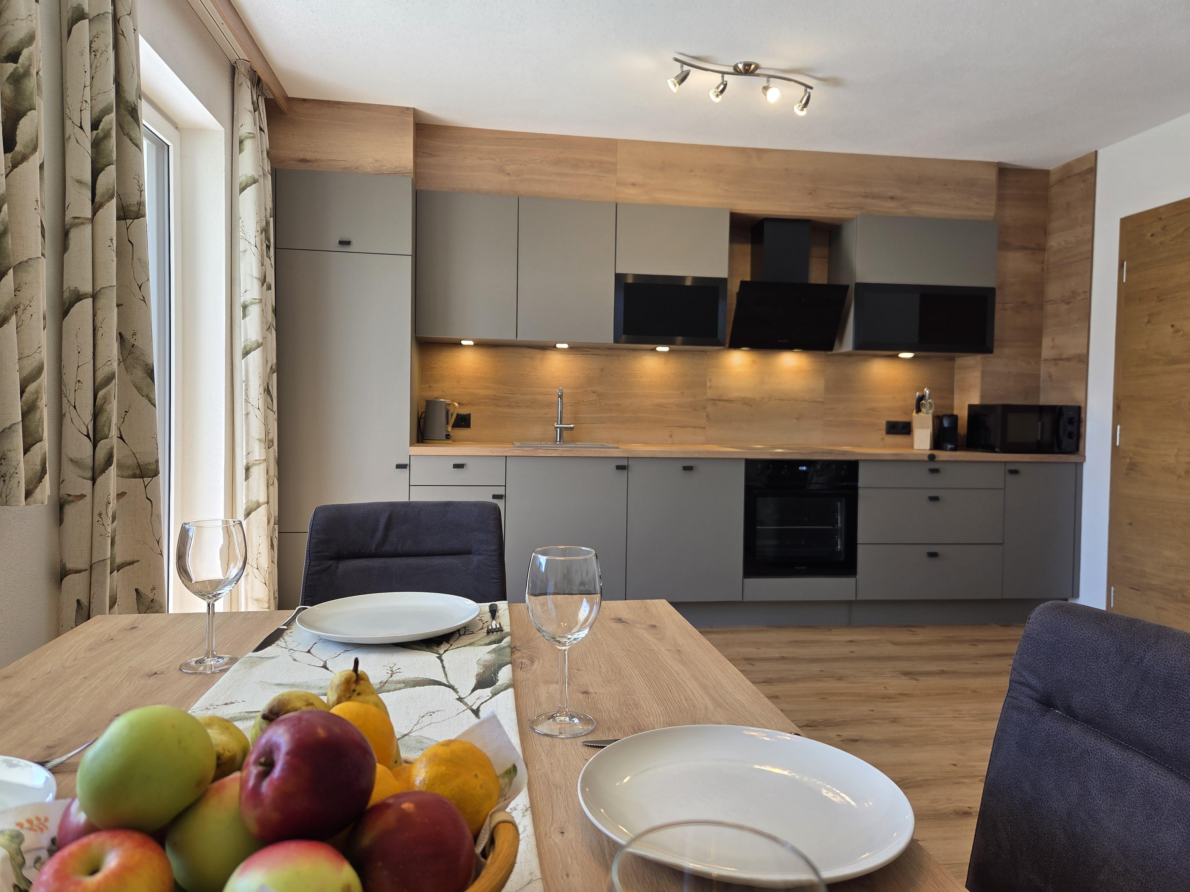 Ein Esstisch mit Obst, leere Teller und Weingläser in einer modernen Küche mit Holz- und Grautönen.
