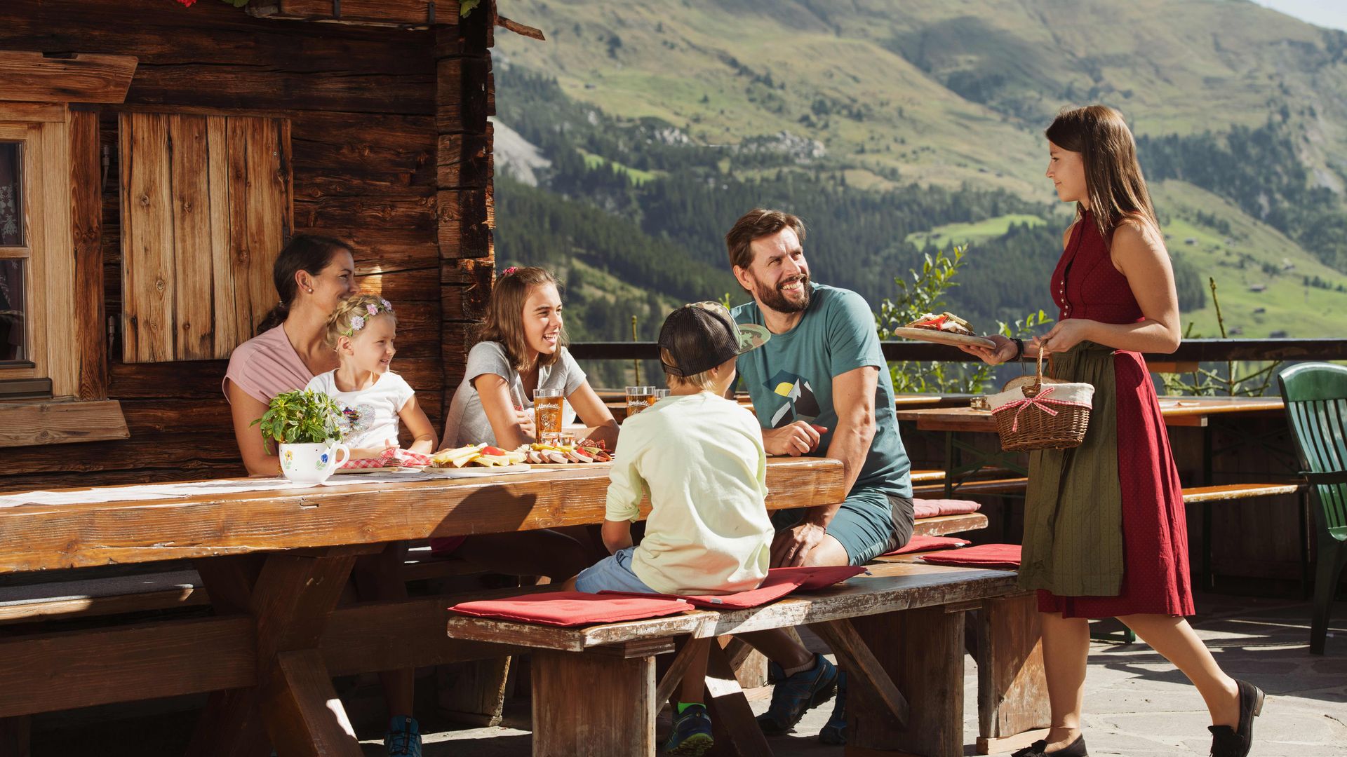 Cherish & Cuisine in Mayrhofen-Hippach Family sitting outside at a wooden table in front of a mountain hut, eating together; woman in traditional clothing serving them. Sunny weather, mountains and nature in the background, friendly atmosphere.