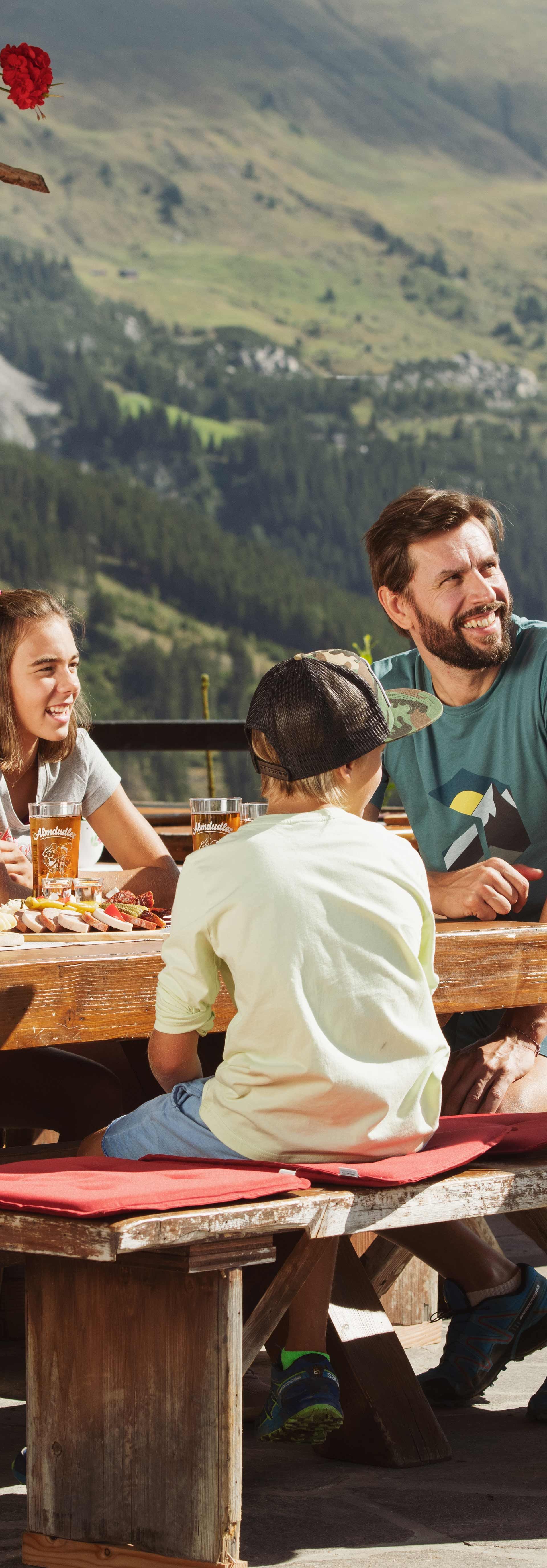 Genuss und Kulinarik in Mayrhofen-Hippach Familie sitzt draußen an Holztisch vor Berghütte und isst gemeinsam; Frau in traditioneller Kleidung serviert. Sonniges Wetter, Berge und Natur im Hintergrund, freundliche Atmosphäre.