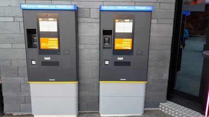 Two self-checkout kiosks with blue signs for purchase, pickup, and return, in front of a stone wall.