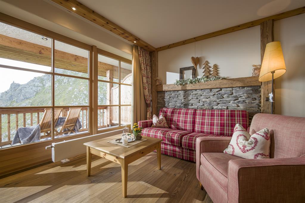 Gemütliches Wohnzimmer mit rotem kariertem Sofa, Sessel, Holzwand, großen Fenstern und Bergblick im Hintergrund.