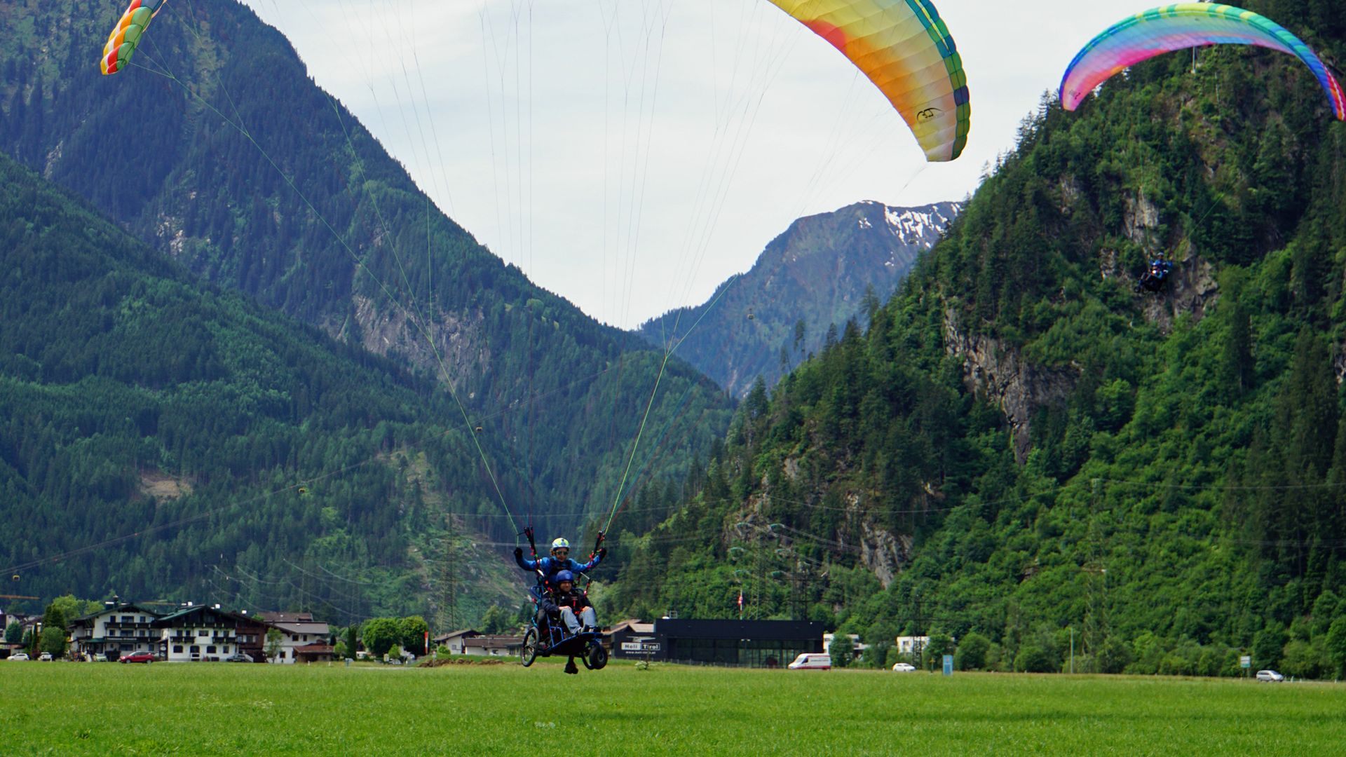 A pilot in wheelchair-supported paragliding lands on a green meadow in Mayrhofen, Zillertal, with a stunning Alpine backdrop.