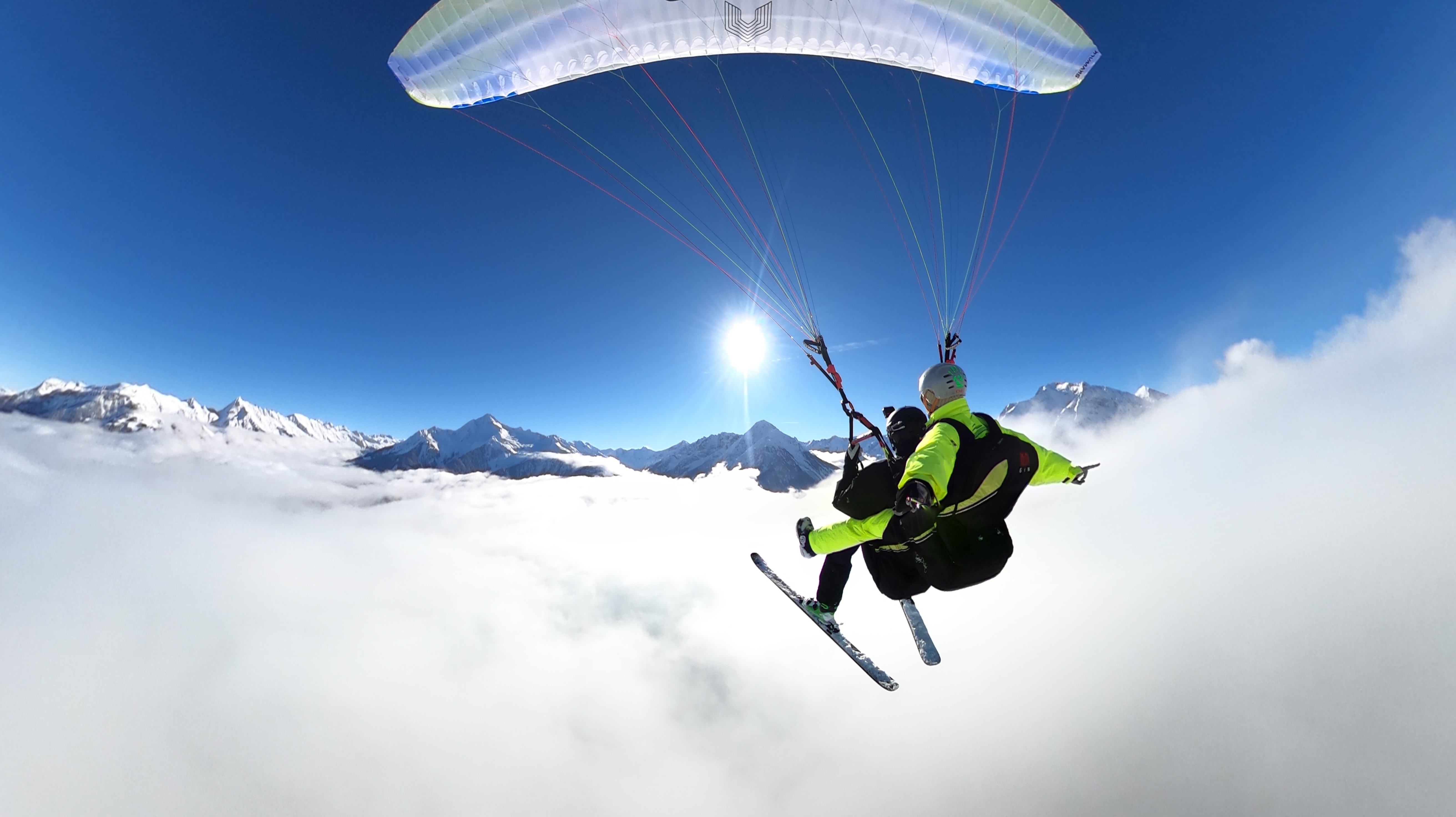 Das Bild zeigt einen Mann mit Skiern an den Füßen, der mit einem Paragleiter hoch über den Wolken fliegt. Er trägt eine Skijacke und Helm und genießt die atemberaubende Aussicht. Der Himmel ist strahlend blau, und unter ihm ziehen weiße Wolken vorbei.