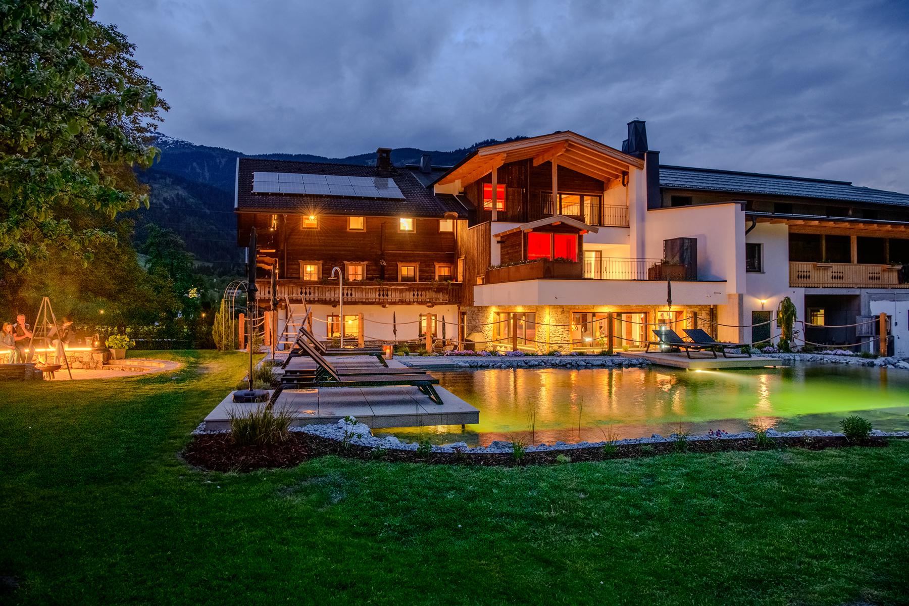 A modern house at night with lit garden, pool, and mountain view in the background.