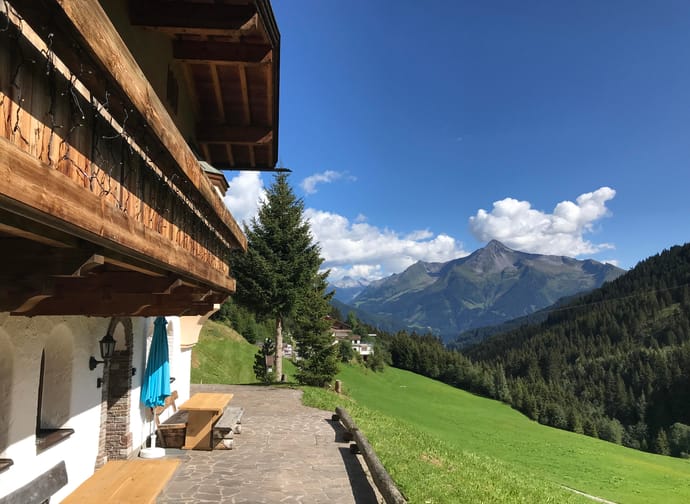 View of a mountain cabin with a wooden terrace, green fields, and mountains under a blue sky.