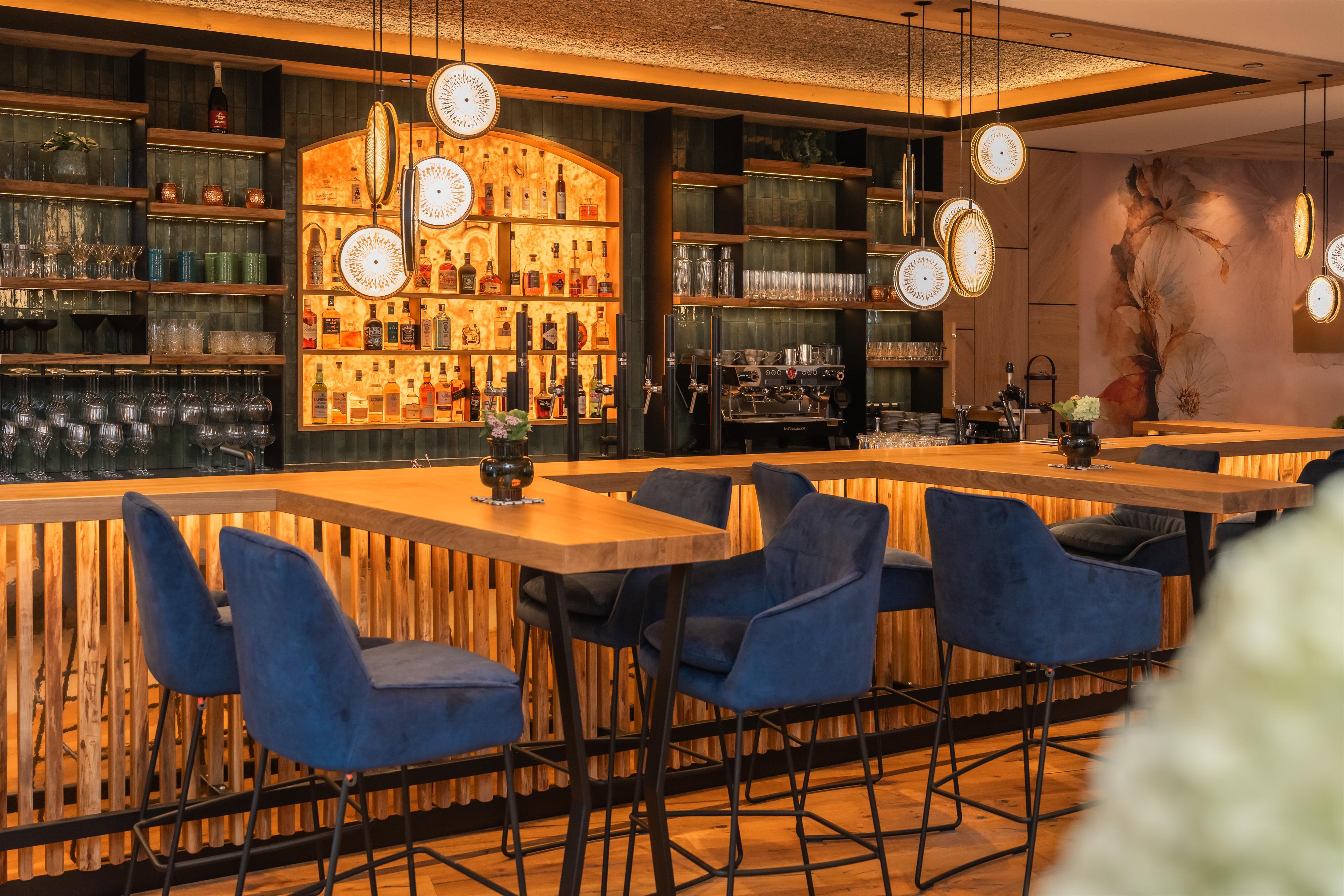 Innenaufnahme einer Bar mit Holzregalen, Gläsern, Flaschen und modernen Hängelampen, warme Beleuchtung, gemütliche Atmosphäre.