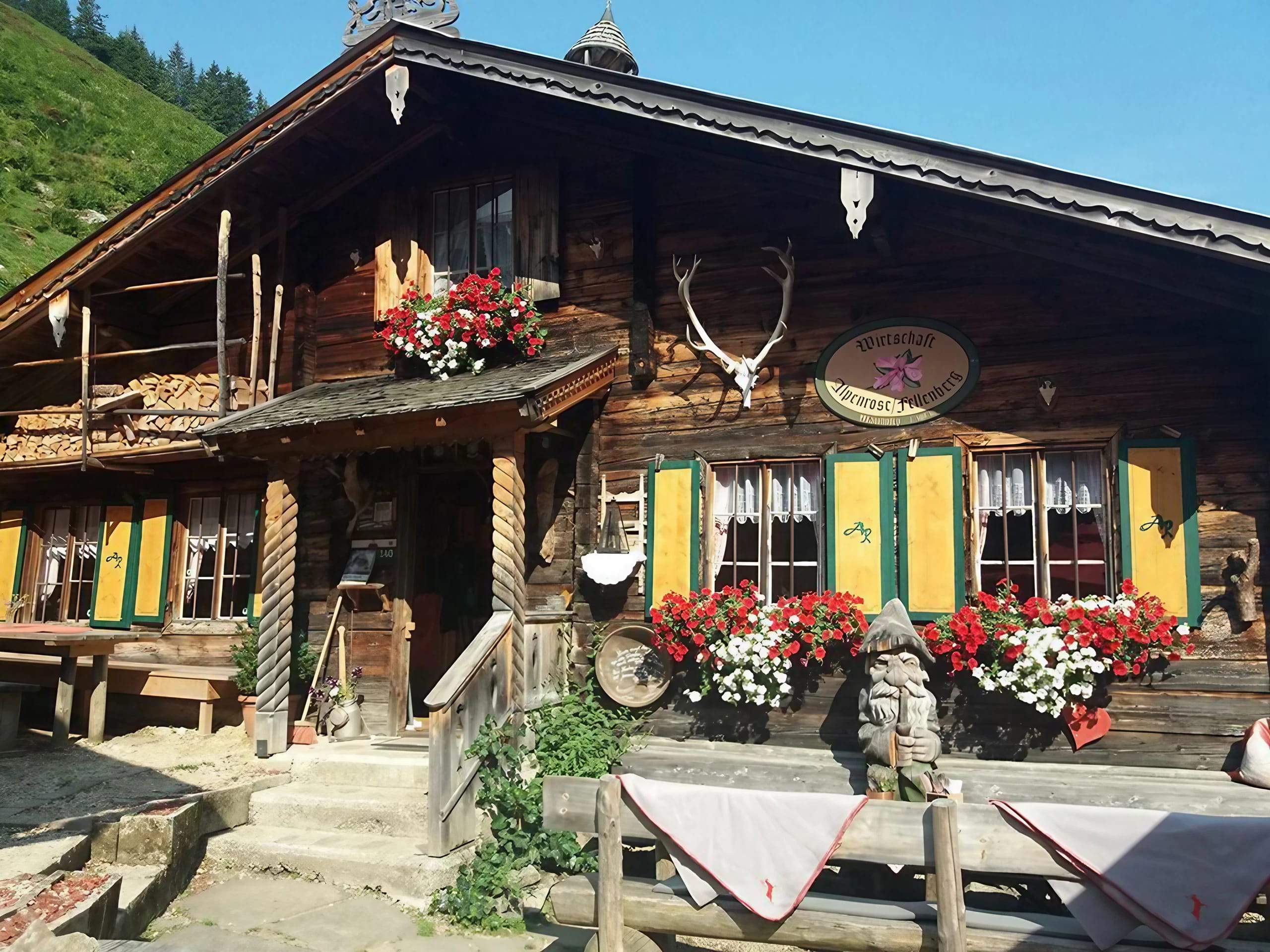 Das Bild zeigt eine rustikale Hütte mit Holzfassade, bunten Fensterläden, Blumendekoration und traditionellen Jagdtrophäen, eingebettet in eine grüne Berglandschaft.