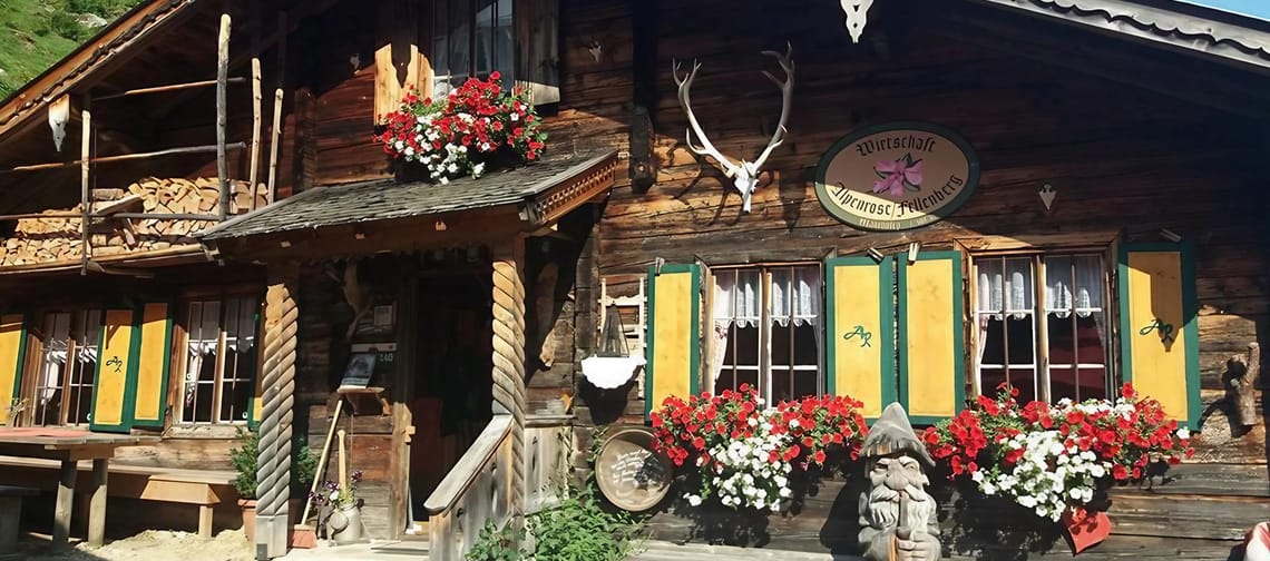 The image shows a rustic mountain hut with a wooden facade, colorful shutters, floral decorations, and traditional hunting trophies, nestled in a green mountain landscape.
