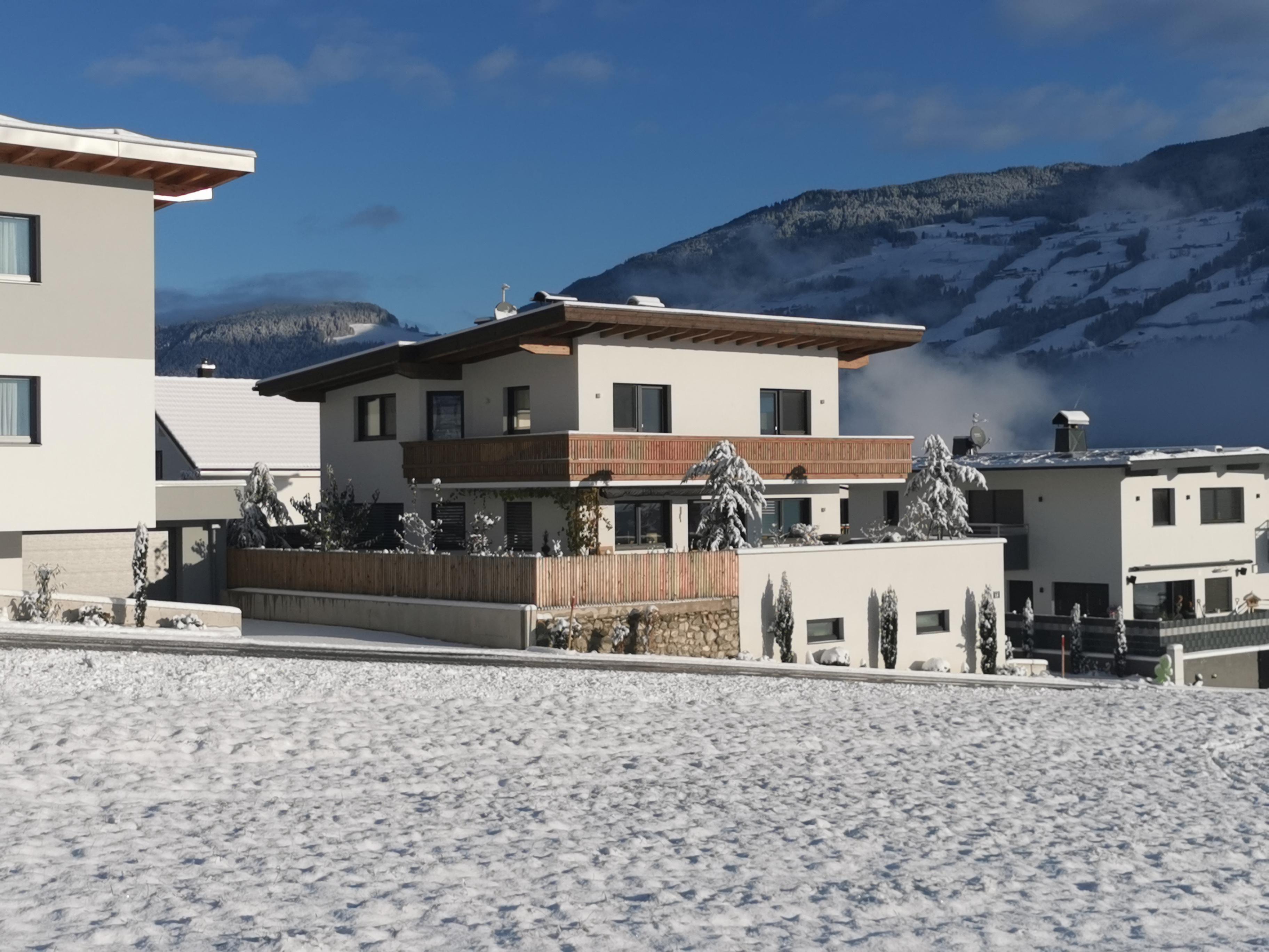 Moderne weiße Häuser mit dunklen Fenstern vor verschneiter Bergkulisse und klarem blauen Himmel.