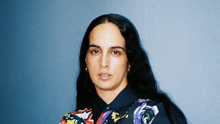 Woman with dark, wavy hair wearing a colorful, artistic shirt against a blue background.