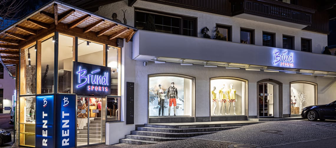 Exterior view of the modern Bründl Sports building in the center of Mayrhofen at night. Large glass facade with lighting, visible logo on the front, wide entrance area at street level.