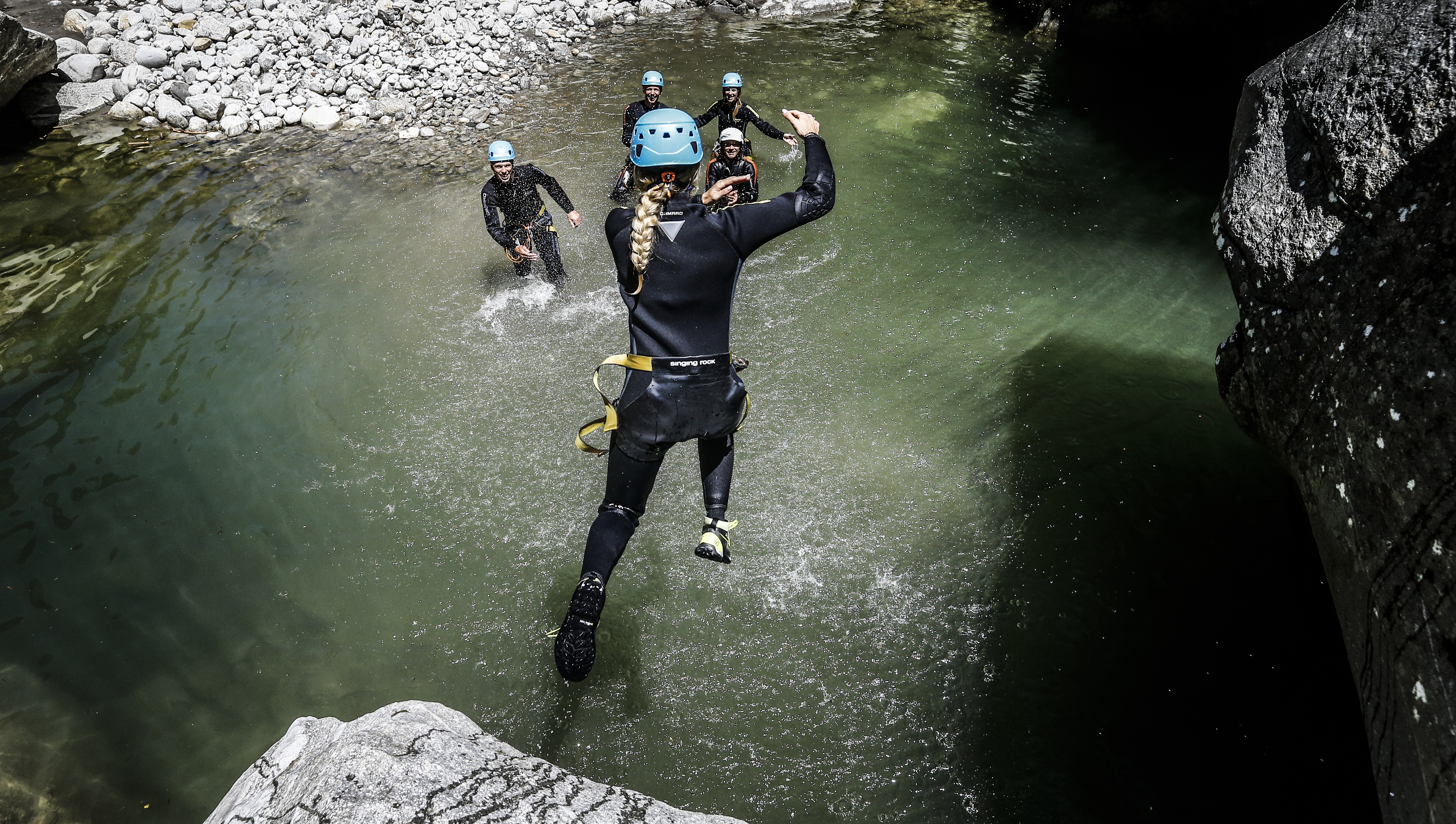 Two people in wetsuits and helmets move through a narrow rocky gorge with clear water – a canyoning scene in an alpine setting.