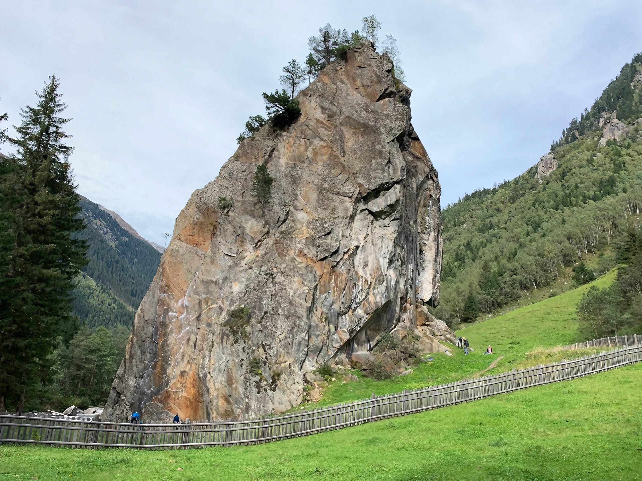 Großer Felsen in grüner Landschaft mit Bäumen, Wiesen und einem Zaun im Vordergrund, bei bewölktem Himmel.