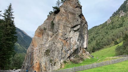 Large rock formation in green landscape with trees, grass, and a fence in the foreground under cloudy sky.