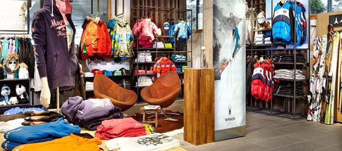 Interior view of a sports store with neatly displayed outdoor and sportswear. Folded jackets and sweaters are laid out on tables, and colorful functional clothing hangs on the walls. Bright, modern store atmosphere.