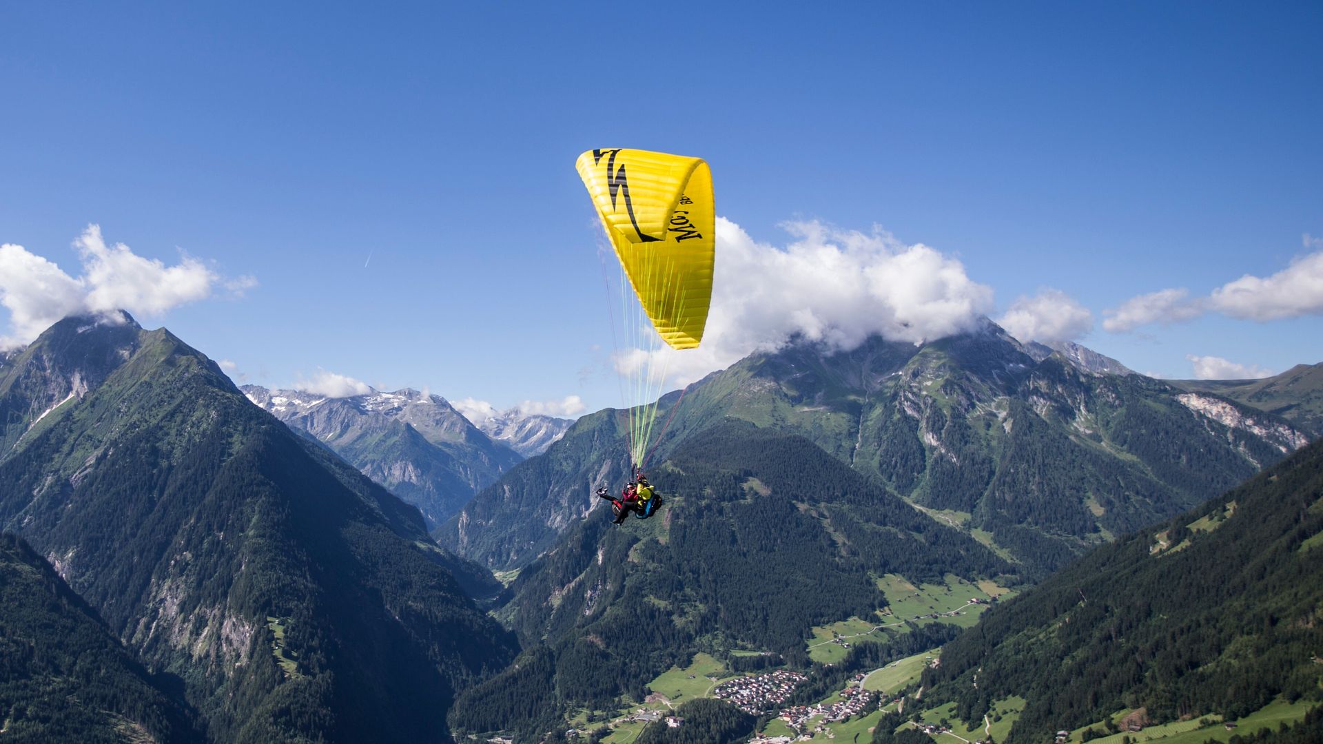 A paraglider with a yellow canopy soars over the Zillertal in Mayrhofen-Hippach. The alpine landscape shines in summer greenery under a blue sky with a few clouds.
