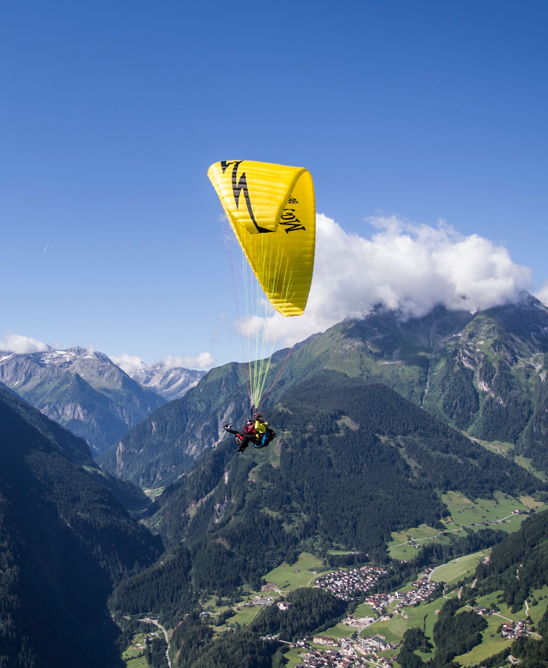 Ein Paragleiter mit gelbem Schirm schwebt über dem Zillertal in Mayrhofen-Hippach. Die Alpenlandschaft leuchtet im sommerlichen Grün unter einem blauen Himmel mit wenigen Wolken.