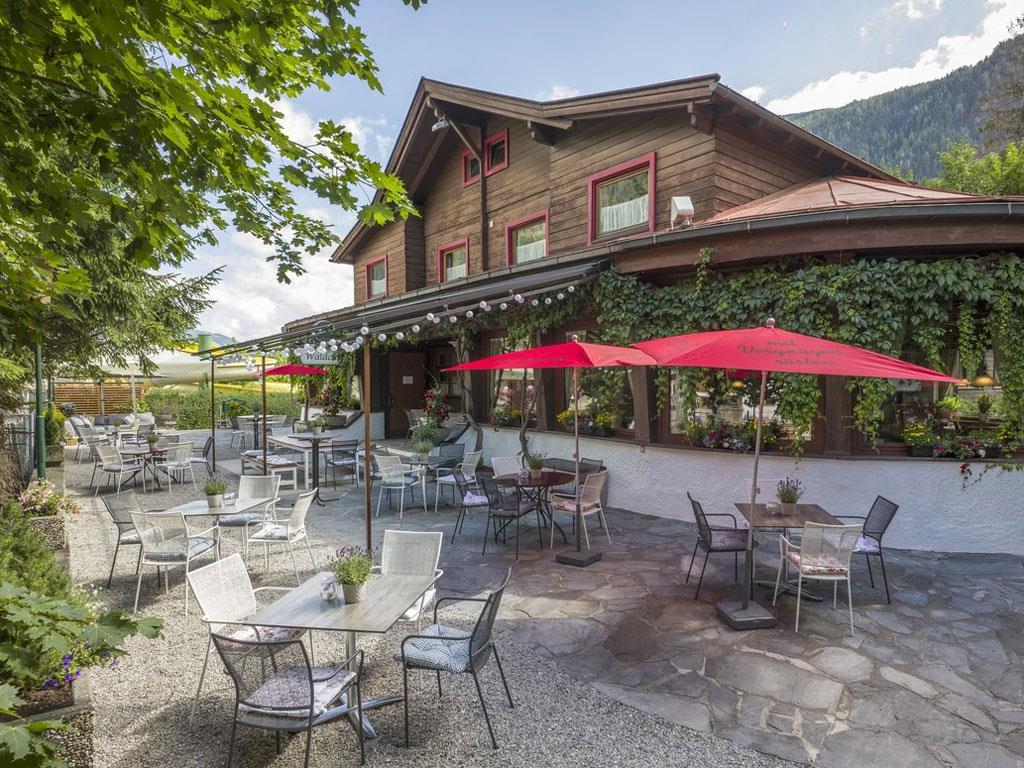 The picture shows Gasthof Waldcafé from the outside. On the terrace, chairs and tables are shaded by red umbrellas, creating a cozy and inviting atmosphere.