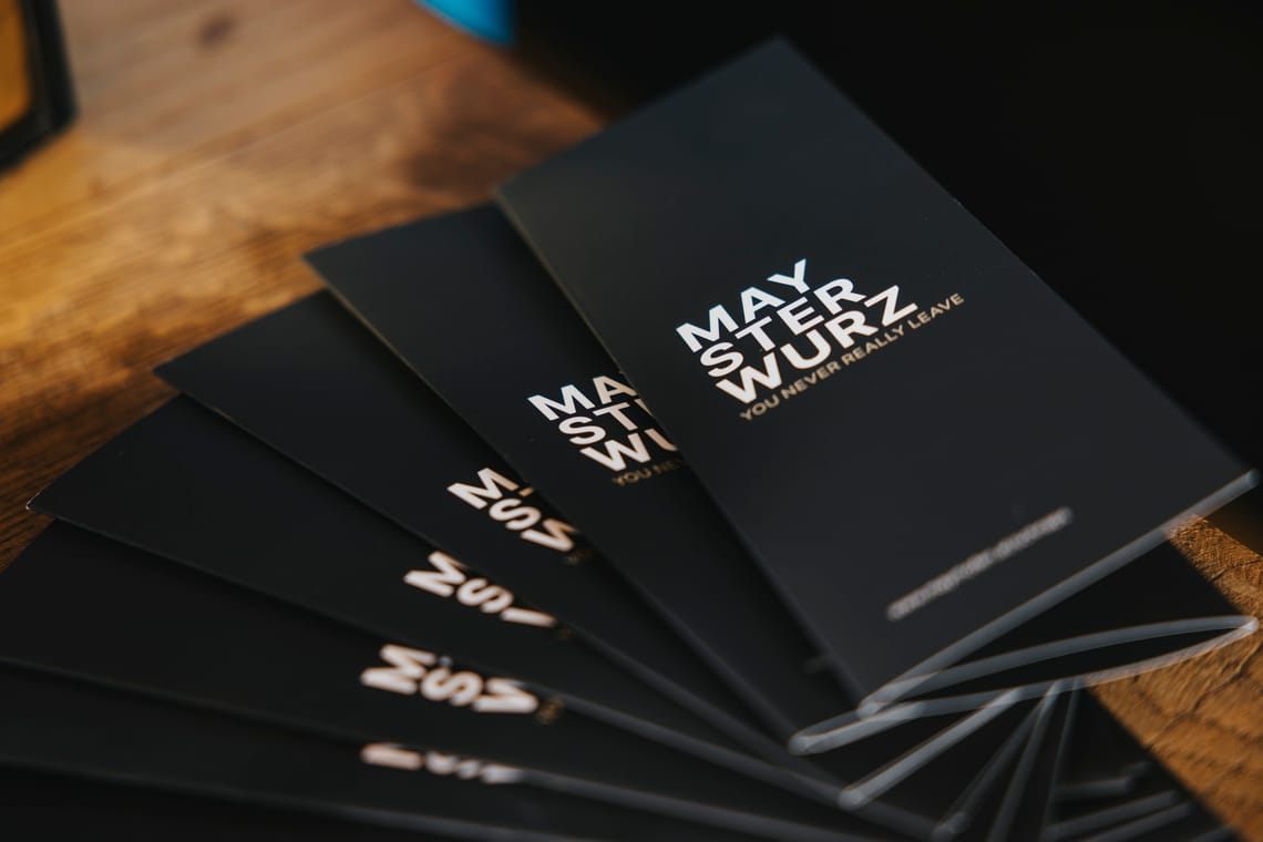 Edelbrand Maysterwurz Schwarze, fächerförmig ausgelegte Flyer mit der Aufschrift „MAYSTERWURZ – VOLL WURZ UND KEIN KRAM“ liegen auf einem Holztisch.