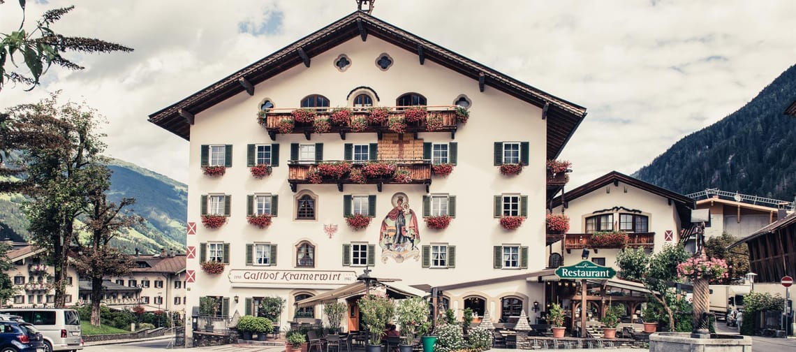 The image shows the inn Kramerwirt, a traditional Alpine building with a white facade, green shutters, and numerous red flower boxes.