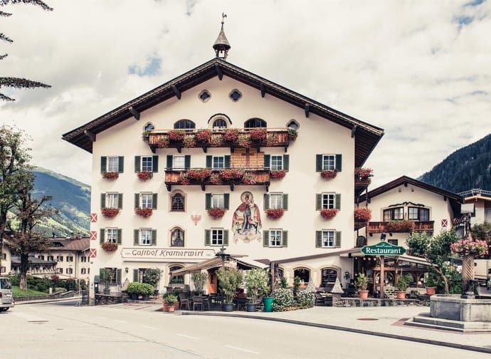 Das Bild zeigt den Gasthof Kramerwirt, ein traditionelles alpenländisches Gebäude mit weißer Fassade, grünen Fensterläden und vielen roten Blumenkästen.